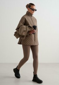 Vadderad brun jacka med hög krage, i kombination med beige leggings och svarta ankelstövlar, håller en kaffekopp, mot en neutral bakgrund.
