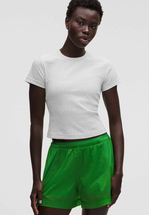 Junge Person mit kurzen lockigen Haaren, die ein schlichtes weißes, eng anliegendes Kurzarmshirt und leuchtend grüne Sportshorts vor hellem Hintergrund trägt.