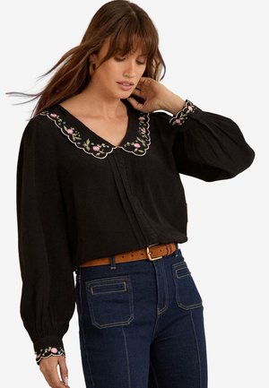 Frau trägt eine schwarze Bluse mit floral besticktem geschwungenem Kragen und Manschetten, kombiniert mit hoch taillierter blauer Jeans und braunem Gürtel.