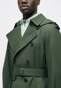 Trench coat verde con design doppiopetto, ampio colletto, cintura in vita e bottoni marroni. Tessuto strutturato con finitura opaca.