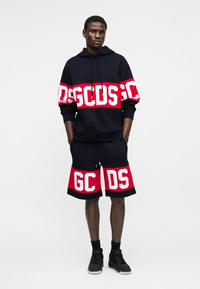 Άνδρας που στέκεται με τα χέρια στις τσέπες, φοράει μαύρο φούτερ με κουκούλα και σορτς με κόκκινα και λευκά γράμματα GCDS, μαύρα αθλητικά παπούτσια και κάλτσες.
