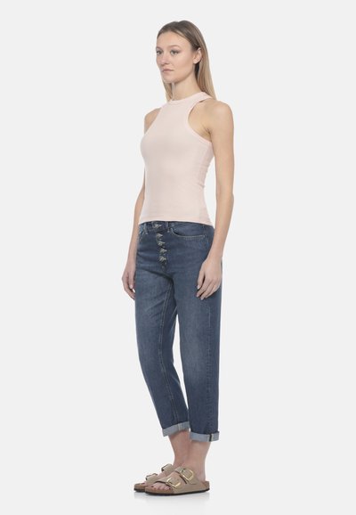 Femme debout portant un jean bleu foncé retroussé avec plusieurs boutons à l'avant, un haut sans manches beige clair, et des sandales beiges à brides avec boucles.