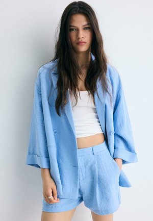 Junge Frau mit langen dunklen Haaren, die einen hellblauen übergroßen Blazer und passende Shorts mit einem weißen, gekürzten Tanktop trägt.