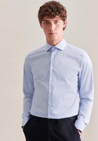 Light blue button-up shirt made of smooth fabric. Features a classic collar, long sleeves, and white buttons. Worn with dark trousers.