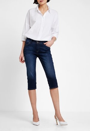 Femme portant une chemise blanche boutonnée, un jean court bleu foncé et des chaussures blanches à talons hauts sur un fond uni.