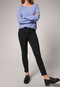 Pull surdimensionné violet clair, jean skinny noir et mocassins noirs épais avec détails dorés, sur un fond neutre.