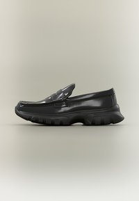 Μαύρος γυαλιστερός δερμάτινος μοκασίνι με χοντρή σόλα, στρογγυλεμένη μύτη και ραμμένα διακοσμητικά στοιχεία πάνω σε απλό ανοιχτό φόντο.