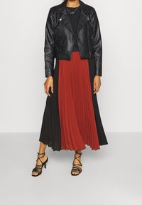 Chaqueta de cuero negra corta sobre una blusa negra y una falda plisada, dividida en secciones rojas y negras. Sandalias de tacón negro con tiras.
