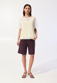 Top senza maniche di un giallo chiaro stratificato sopra una camicia bianca trasparente a maniche corte, abbinato a pantaloni corti viola scuro e sandali con cinturini.