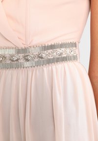 Robe rose pâle avec une jupe texturée plissée et une ceinture argentée ornée de pierres précieuses, présentant des formes de diamant et des accents métalliques.