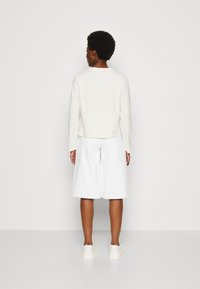 Felpa bianca a maniche lunghe, design cropped; abbinata a pantaloni corti bianchi a gamba larga. Sneaker bianche piatte, estetica semplice e pulita.