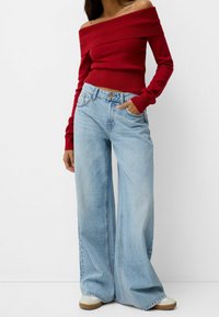 Femme portant un pull rouge à épaules dénudées, un jean large bleu clair et des baskets blanches, une main dans la poche, sur un fond uni.