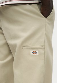 Μπεζ παντελόνια Dickies με πλαϊνή τσέπη και λογότυπο ραμμένο στο ύφασμα· χέρι που βουτάει στην μπροστινή τσέπη, εμφανίζοντας υφή υφάσματος και λεπτομέρειες ραφής.