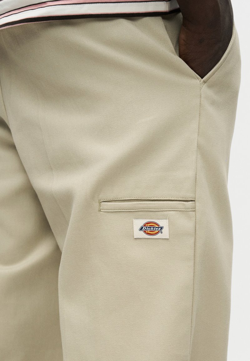 Pantalon beige Dickies avec une poche latérale et un patch logo ; main glissée dans la poche avant, mettant en valeur la texture du tissu et les détails de la couture.
