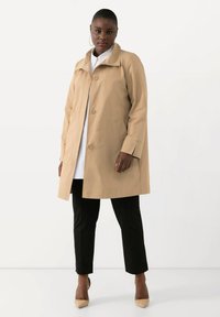Cappotto trench impermeabile beige con colletto, chiusura a singolo bottone, due tasche laterali e maniche lunghe, indossato sopra una camicia bianca e pantaloni neri.