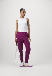 Witte geribbelde tanktop met zwart Nike-logo, paarse geplooide broek, houdt een witte tennisracket vast, draagt witte en paarse sportschoenen.