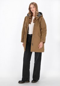Bruine parka jas met een grijze capuchon, voorzien van een rits aan de voorkant en drukknoopsluitingen, zijzakken en een relaxte pasvorm, gecombineerd met zwarte broek en laarzen.