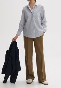 Gestreifte Button-Down-Bluse in Weiß und Marineblau, lange Ärmel, lässige Passform. Kombiniert mit hellbraunen, weit geschnittenen Hosen und einer dunklen Jacke. Weiße Sneakers.