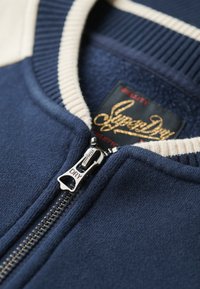 Granatowa kurtka na suwak z teksturowanej tkaniny, kremowym prążkowanym kołnierzem, srebrnymi detalami zamka oraz czarną metką z złotymi literami.