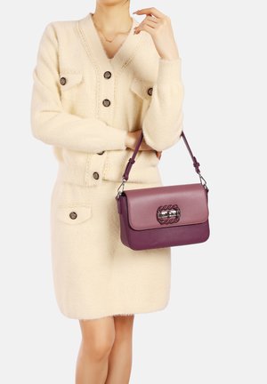 Vestito cardigan in pelliccia color crema con bottoni e tasche, abbinato a una borsa a due tonalità di viola con dettagli in catena e tracolla regolabile.