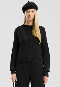 Cardigan en tricot noir avec un col en V, fermeture à boutons et manches longues. Porté sur un haut noir côtelé, assorti à un pantalon noir.