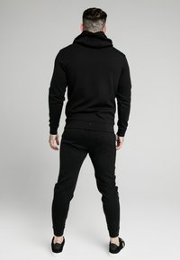 Set di felpa nera con cappuccio e pantaloni jogger in materiale morbido, con una vestibilità comoda, orli delle caviglie elastici e un grande cappuccio. Dettaglio logo discreto sui pantaloni jogger.