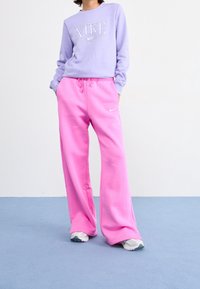 Fioletowa bluza z długim rękawem z logo "NIKE", zestawiona z luźnymi różowymi spodniami dresowymi. Posiada elastyczny pas i klasyczne trampki.