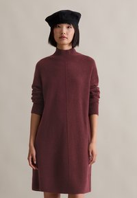 Frau mit glattem schwarzen Haar, die eine schwarze Baskenmütze und ein langärmeliges, kastanienbraunes Strickkleid vor einem schlichten beige Hintergrund trägt.