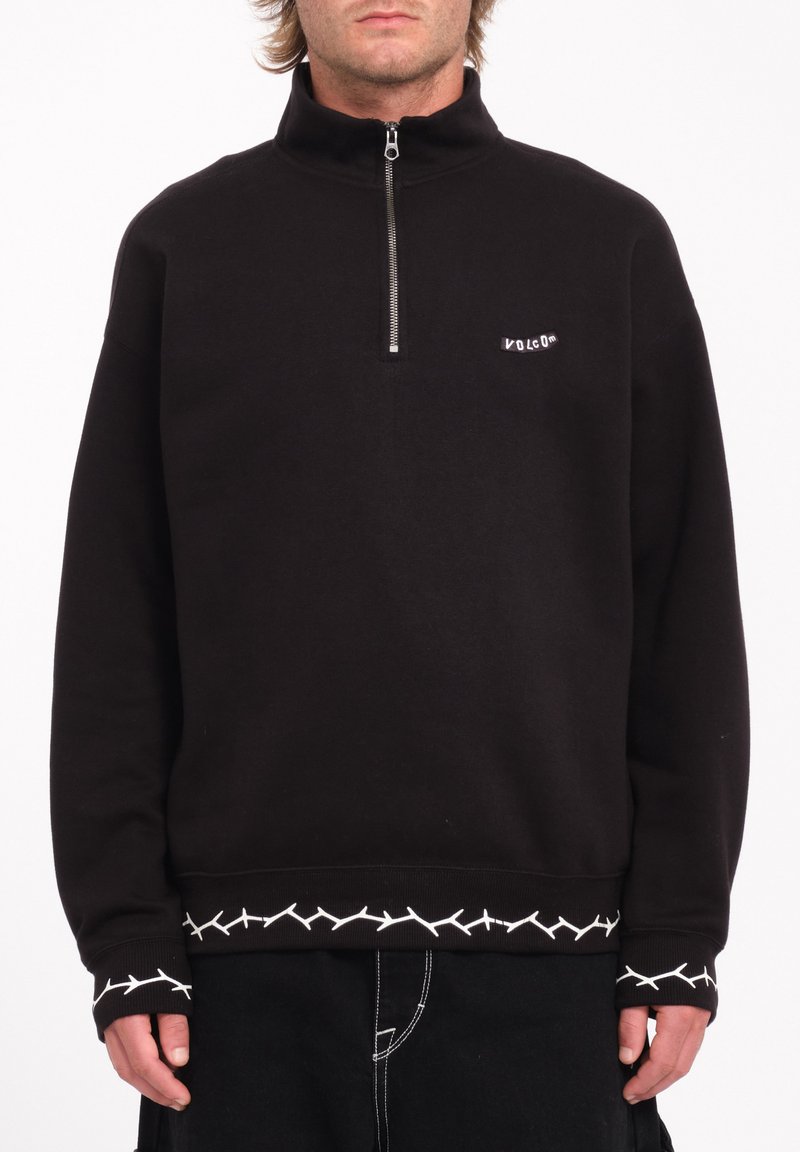 Sudadera negra con cremallera y cuello alto, con acentos de costura blanca en el dobladillo y los puños, y un pequeño logo en el pecho.