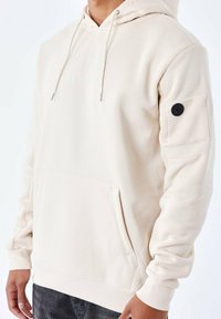 Sudadera crema con un bolsillo canguro en el frente, cordones y un acento circular negro en la manga. Tejido suave de mezcla de algodón; corte holgado.