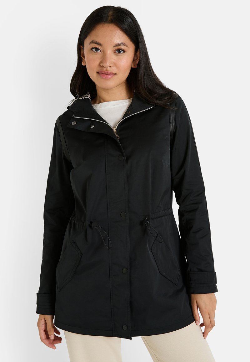 Mujer con cabello largo y oscuro, vistiendo una chaqueta negra con cremallera y cordones en la cintura, y pantalones beige, frente a un fondo blanco.