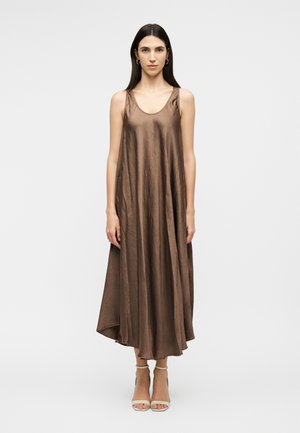 Kvinde med langt mørkt hår iført en ærmeløs, løstsiddende brun midi-kjole og hvide ankelremssandaler, stående foran en ensfarvet baggrund.