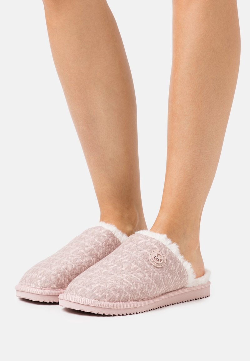 Pieds portant des chaussons à enfiler rose clair à motifs, avec une doublure en fourrure blanche et un petit logo circulaire sur le côté.