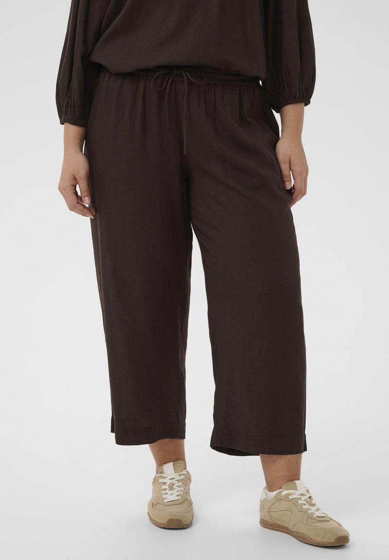 Personne portant un pantalon ample marron foncé coupé court avec une taille élastique et des baskets beige, debout devant un fond uni.