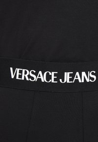 Black ribbed fabric with a black elastic waistband featuring "VERSACE JEANS" in bold white letters. Smooth texture, fitted design.