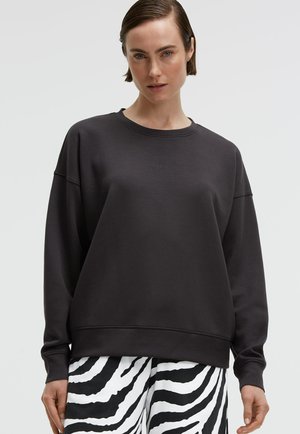 Frau trägt einen schlichten, schwarzen, übergroßen Pullover und schwarz-weiße Zebra-Musterhose, posiert vor neutralem Hintergrund.