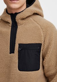Brun fleece pullover med en sort lynlåslomme på brystet. Har en hætte, tekstureret stof og en delvis lynlåslukning ved halsen.