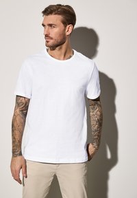 Bílý bavlněný tričko s kulatým výstřihem a ohrnutými krátkými rukávy, které vyniká hladkým povrchem. Model nosí béžové kalhoty s viditelnými tetováními.