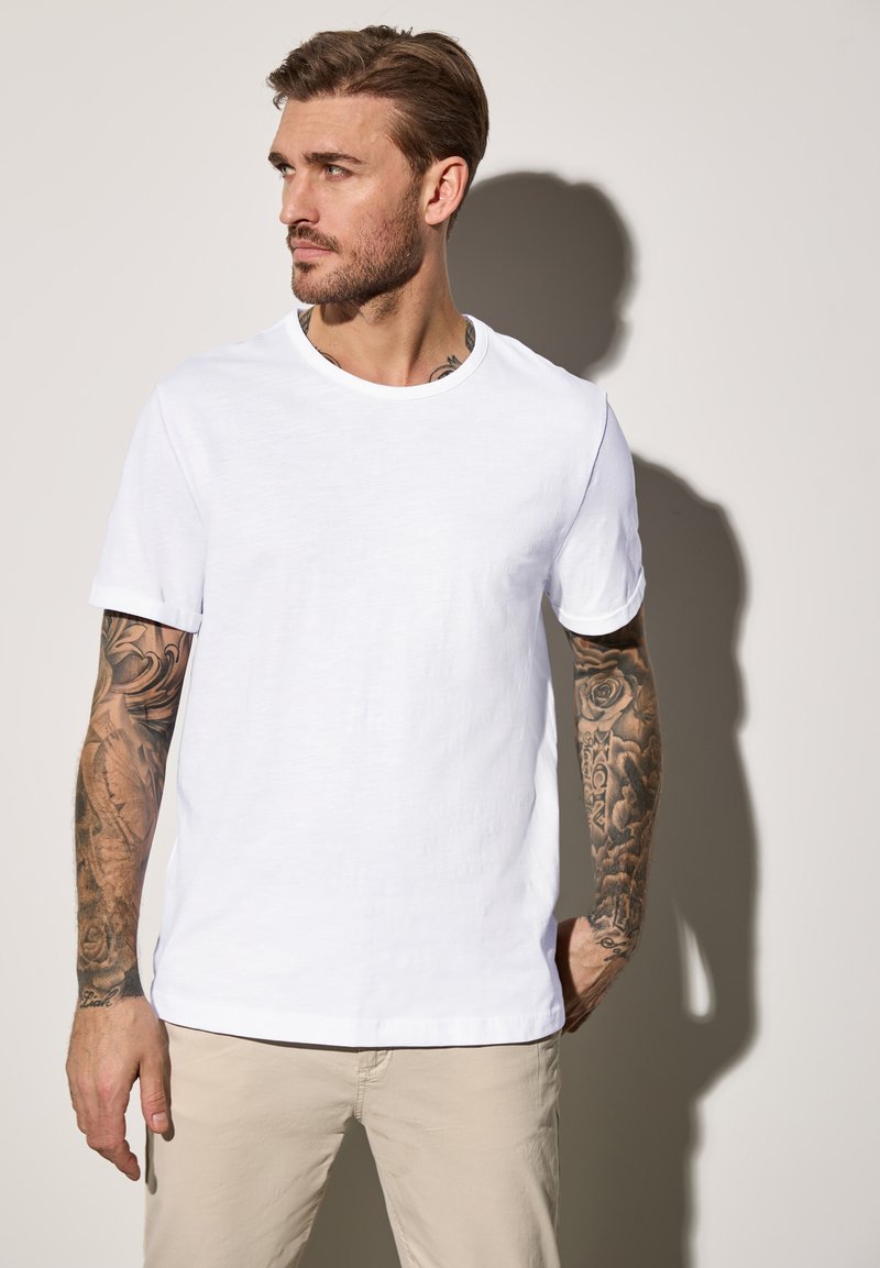 Bílý bavlněný tričko s kulatým výstřihem a ohrnutými krátkými rukávy, které vyniká hladkým povrchem. Model nosí béžové kalhoty s viditelnými tetováními.