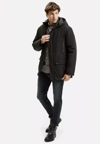 Mężczyzna w czarnej kurtce z kapturem, w koszulę w kratę, ciemne dżinsy i czarne buty, stoi z jedną nogą skrzyżowaną i patrzy na bok.