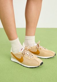 Beiga träningsskor med orange Nike-swoosh, texturerad ovandel och vita snörningar. Bärs med ljusa ankelsockor på grön yta.