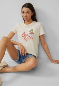 Crème-kleurige t-shirt met een "Sea La Vie" graphic in koraal, blauw en groen, gecombineerd met denim shorts en beige platform sandalen.