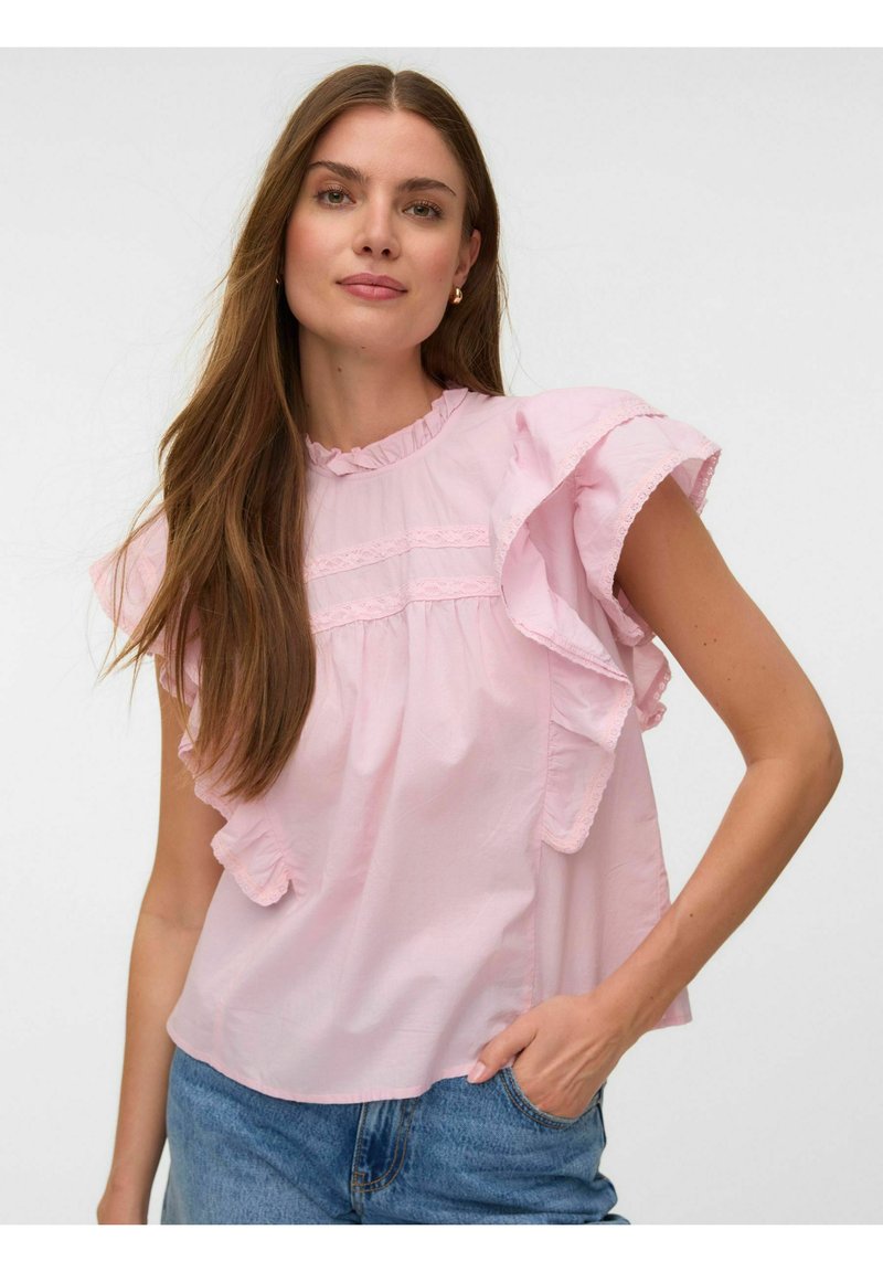 Femme aux longs cheveux bruns portant un chemisier rose clair à volants et un jean bleu, debout avec une main dans la poche sur fond blanc.