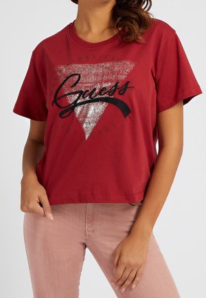 Rode katoenen T-shirt met een cropped ontwerp. Heeft een glinsterende driehoekige grafiek en zwarte tekst "Guess." Korte mouwen en een ronde halslijn.