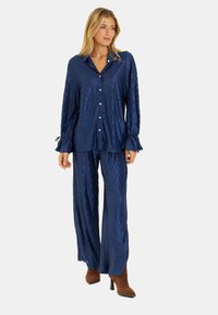 Granatowy satynowy zestaw piżamowy z teksturowanym wzorem w diamenty, zapinana na guziki koszula z falbanami i pasujące szerokie spodnie. Brązowe botki na obcasie.