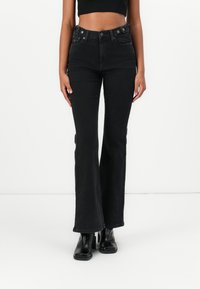 Black flared jeans in denim with a high waist, featuring metal button accents and side pockets. Paired with black ankle boots.