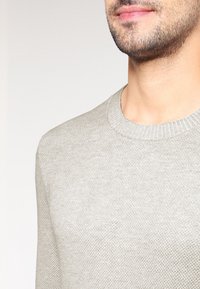 Pull en maille gris clair avec col rond, tissu texturé, poignets côtelés et finition lisse, montrant un gros plan de la partie supérieure de la poitrine.