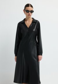 Žena v dlouhé černé kožené šaty s knoflíky a límečkem, černými slunečními brýlemi a stříbrnými kruhovými náušnicemi na bílém pozadí.