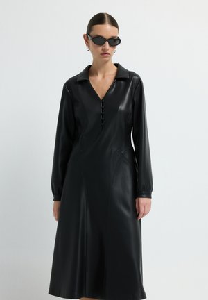 Vrouw in een lange zwarte leren jurk met knopen en kraag, zwarte zonnebril en zilveren creolen tegen een witte achtergrond.