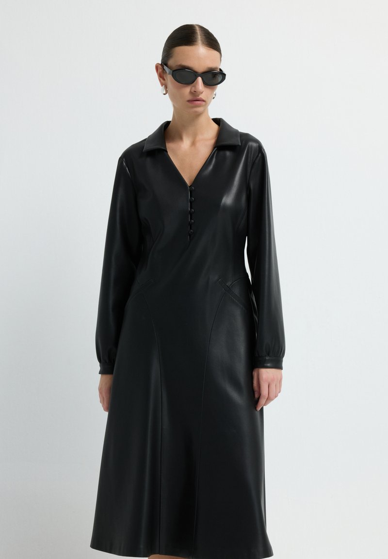 Žena v dlouhé černé kožené šaty s knoflíky a límečkem, černými slunečními brýlemi a stříbrnými kruhovými náušnicemi na bílém pozadí.